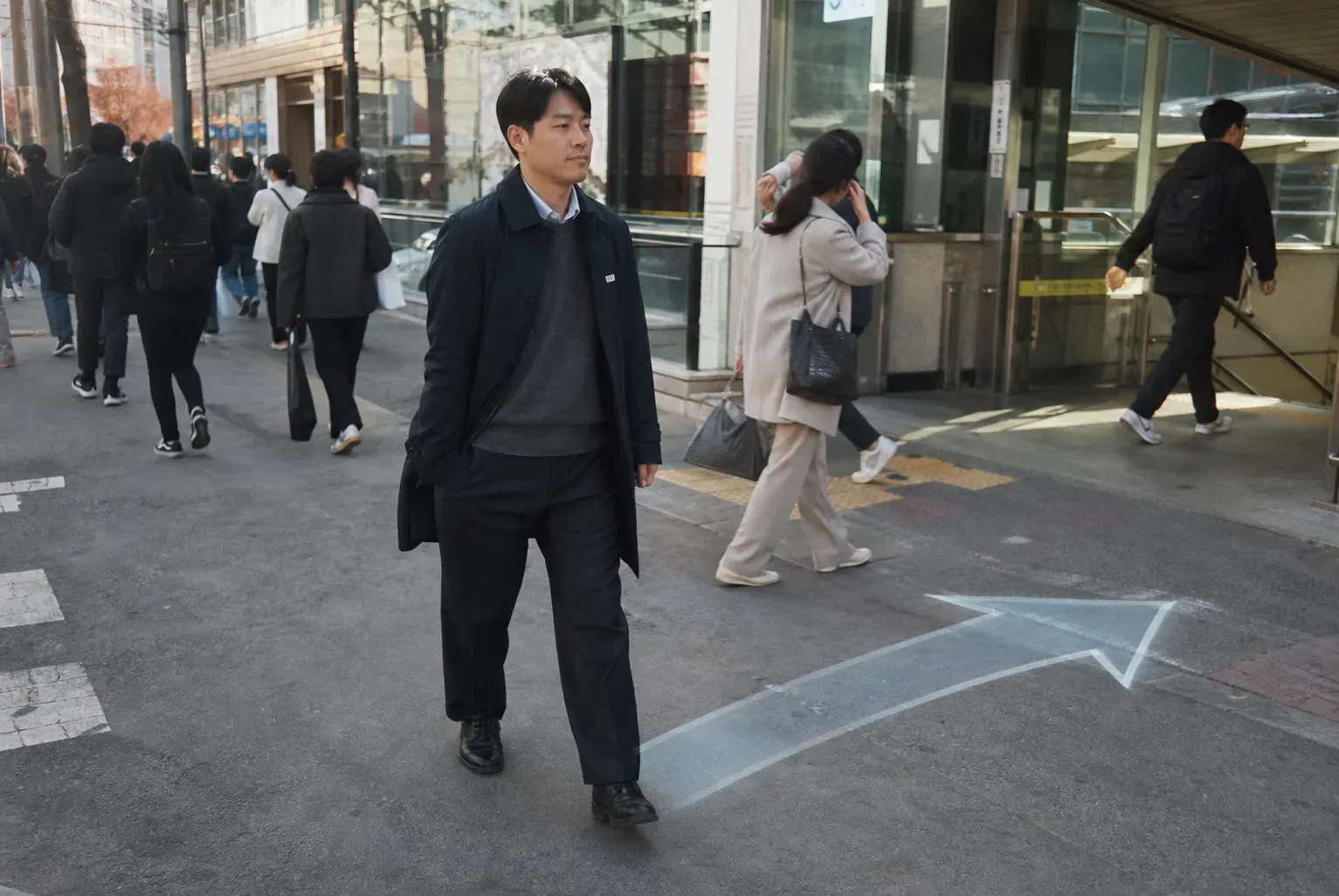 서울 성수동 지하철 출구를 나온 이진우가 스마트폰을 꺼내지 않은 채 시야 가장자리의 길안내 화살표를 바라보며 걷고 있고 주변 시민들은 그를 평범한 직장인처럼 지나치는 장면